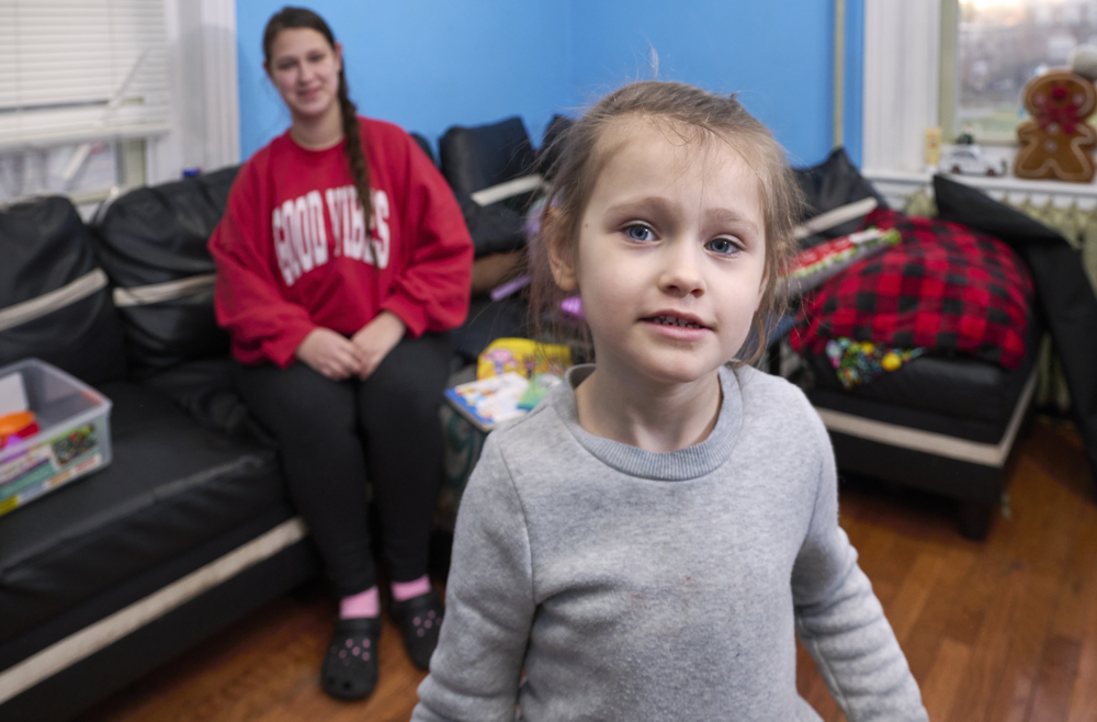 Scarlett Adams, a five-year-old, looks into the camera, with her mother sitting on a couch behind her.